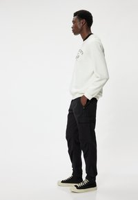 Weißer Sweatshirt mit schwarzer Schrift, kombiniert mit schwarzen Cargo-Hosen mit seitlichen Taschen. Schwarze High-Top-Sneaker mit cremefarbenen Sohlen.