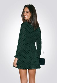 Vestido verde oscuro con lunares negros, dobladillo con volantes, cintura fruncida y mangas largas. Combinado con un pequeño bolso negro.