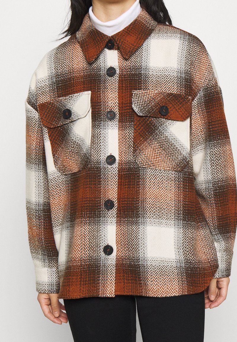 Femme portant une veste à carreaux marron, blanc et gris avec des boutons et de grandes poches avant sur un col roulé blanc.