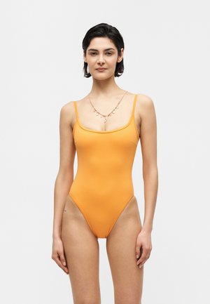 Kobieta z krótkimi ciemnymi włosami, ubrana w pomarańczowy strój kąpielowy bez rękawów i złoty naszyjnik, stojąca na jednolitym białym tle.