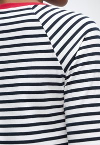 Κοντινό πλάνο ενός ατόμου που φοράει λευκό πουκάμισο με οριζόντιες μαύρες ρίγες σε ίσες αποστάσεις και κόκκινο λαιμόκοψη, δείχνοντας τον ώμο και το πάνω μέρος του μπράτσου.