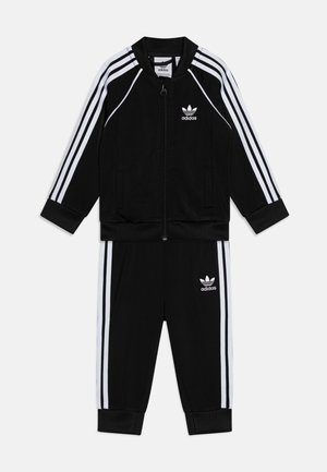 Fato de treino preto da adidas com design de três riscas brancas, apresentando uma jaqueta com fecho éclair e punhos canelados, juntamente com calças combinadas. Logótipo em ambas as peças.