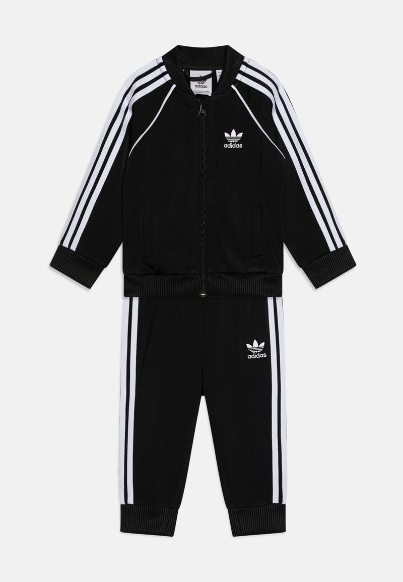Svart adidas träningsoverall med vit tre-randig design, bestående av en zipjacka med ribbade muddar och matchande byxor. Logotyp på båda plaggen.
