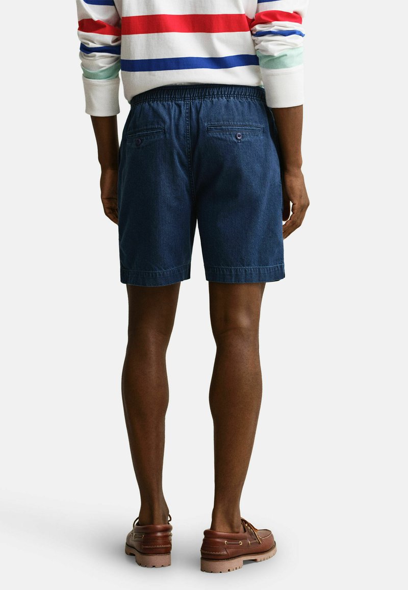 Persona che indossa pantaloncini di jeans blu, una camicia bianca a maniche lunghe con strisce rosse e blu, e scarpe da barca in pelle marrone, vista di spalle.