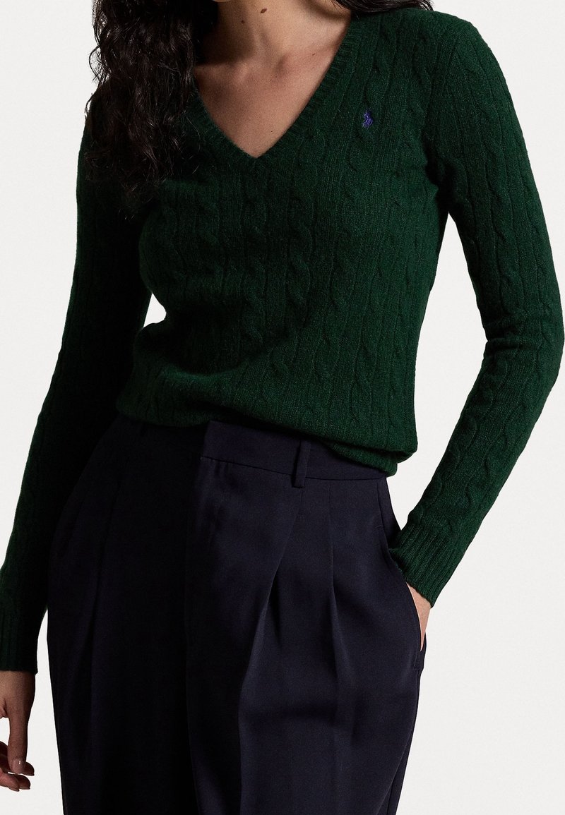 Femme portant un pull à col en V en maille torsadée vert foncé et un pantalon noir plissé taille haute, main dans la poche.
