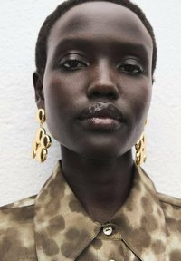 Gold drop earrings with circular shapes, worn with a light brown, patterned shirt featuring a collar. Smooth texture and polished finish visible.