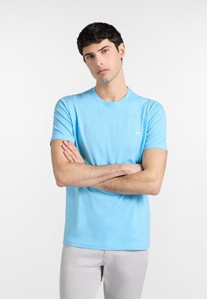 Mladý muž s tmavými kučeravými vlasmi, oblečený v svetlomoddedom tričku a svetlosivých nohaviciach, stojaci so založenými rukami na bielom pozadí.