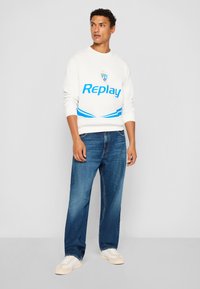 Hvid sweatshirt med blå "Replay" logo og detaljer, parret med mørkeblå jeans og lyse sneakers. Model står imod en ensfarvet baggrund.