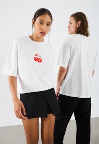 Hvide kortærmede t-shirts har et rødt kirsebærgrafik foran. Modellen bærer en sort plisseret nederdel, mens en anden model bærer sorte bukser.
