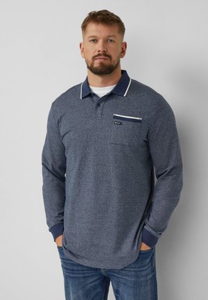 Homme à la barbe taillée portant un polo bleu marine à manches longues avec une poche poitrine et un jean, debout devant un fond gris uni.