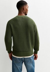 Maglione verde lavorato a maglia con una texture a coste, spalle ripiegate e scollatura ampia; abbinato a jeans blu chiaro. Vestibilità su misura, design semplice.