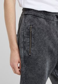 Närbild av en person som bär mörkgrå tvättade byxor med elastisk midja och en vertikal dragkedjeficka på sidan.