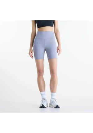 Femme portant un cuissard taille haute violet clair, des chaussettes blanches, des chaussures de sport blanches et un crop top noir, debout devant un fond blanc uni.