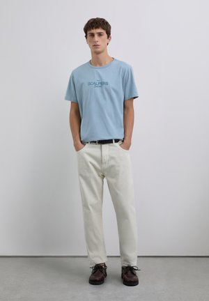 Junger Mann steht mit den Händen in den Taschen, trägt ein hellblaues T-Shirt mit Logo, cremefarbene Hosen, einen schwarzen Gürtel und dunkelbraune Schuhe.