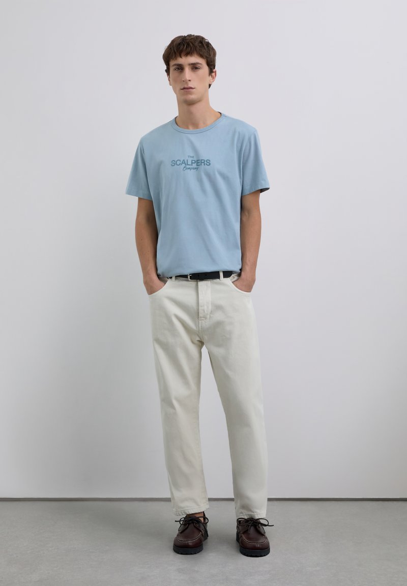 Joven de pie con las manos en los bolsillos, vistiendo una camiseta de logo azul claro, pantalones color crema, cinturón negro y zapatos marrón oscuro.