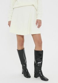 Witte pluizige rok met een subtiele textuur, gecombineerd met kniehoge glanzende zwarte laarzen met een blokhak. Eenvoudig, strak ontwerp.