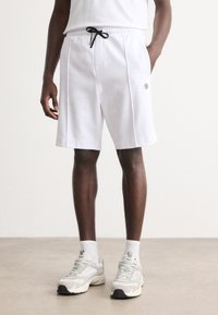 Shorts en polaire blanche avec une taille élastique et un cordon de serrage noir, présentant un détail de logo sur le côté et une coupe décontractée.