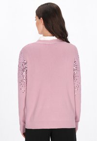 Svetle růžový svetr s žebrovanou texturou, vyznačující se flitrovými ozdobami na ramenou. Klasický límeček a uvolněný střih.