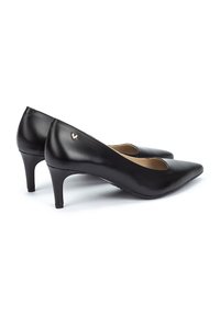 Zapatos de tacón alto de cuero negro con puntera afilada, corte lateral y un tacón moderado. Presentan un pequeño acento de logo dorado en la parte posterior.