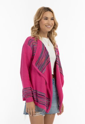 Donna sorridente che indossa un cardigan rosa acceso lavorato a maglia con righe multicolori sopra una maglietta bianca e una gonna di jeans con orlo sfrangiato.