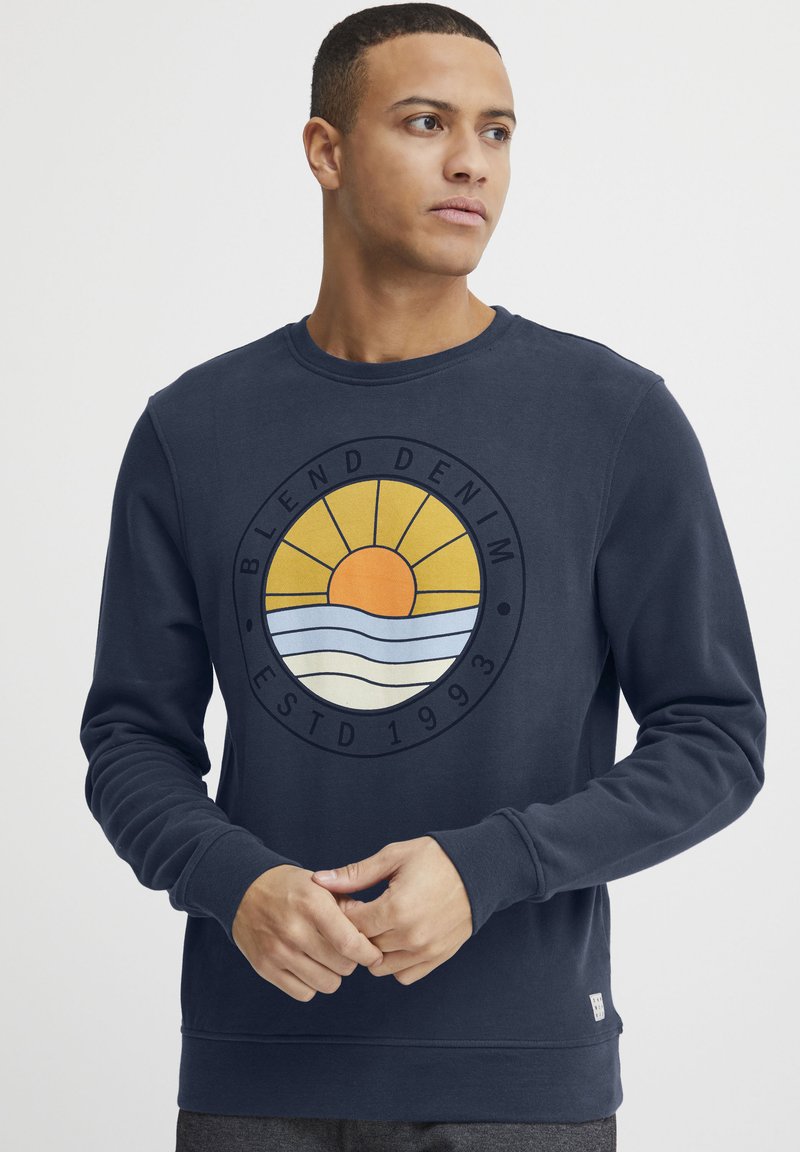 Jūrinės spalvos prigludęs džemperis su apvaliu, daugiakryptiu grafinu, kuriame vaizduojama saulė, bangos ir tekstas. Raibi rankogaliai ir apatinė dalis, medvilnės ir sintetinių pluoštų mišinys.