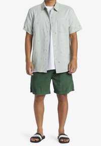 Camicia verde chiaro a maniche corte con taschino e motivi floreali multicolori, abbinata a pantaloni corti in velluto a coste verde scuro e sandali neri.