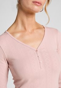 Close-up van een vrouw die een lichtroze textuurtop met een V-hals en drie kleine knopen draagt, waarbij haar hals en een deel van haar gezicht zichtbaar zijn.