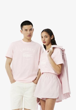 T-shirt in cotone rosa chiaro con logo bianco Lacoste; abbinato a pantaloni bianchi e una gonna a righe rosa. Il modello tiene un giubbotto a righe.