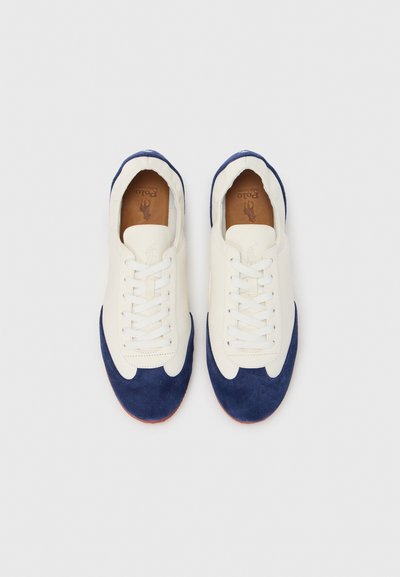 Fehér vászon tornacipő sötétkék velúr részletekkel, lekerekített orral, fűzős bekapcsolódással és világosbarna belső béléssel.