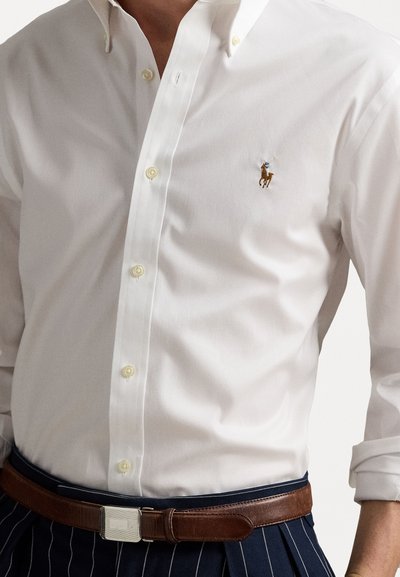 Chemise blanche à manches longues en coton, ornée d'un petit logo brodé marron sur la poitrine gauche, associée à une ceinture en cuir marron et un pantalon à rayures.