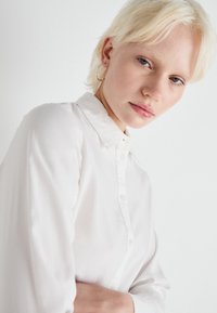 Camicia bianca con bottoni, dal tessuto morbido e liscio, e colletto classico. Il modello ha i capelli biondi corti e indossa orecchini a cerchio in argento.