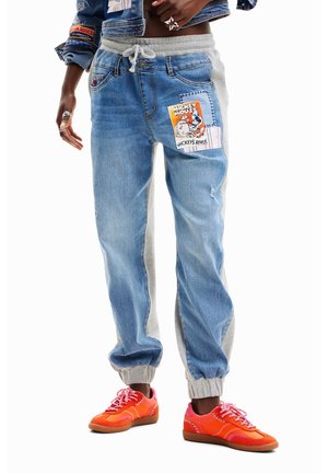 Person iført lyseblå denim og grå joggebukser med Mickey Mouse-patch samt lyseorange sneakers med lyserøde snørebånd.