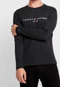 Schwarzes langärmliges Shirt mit Rundhalsausschnitt, versehen mit dem weißen "TOMMY HILFIGER" Logo und dem Text "EST. 1985". Weicher Stoff mit eng anliegenden Ärmeln.