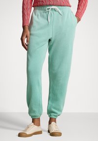 Světle azurové teplákové kalhoty s elastickým pasem, bílou tkanicí, manžetovými nohavicemi a měkkou texturou. V kombinaci se krémovými teniskami.