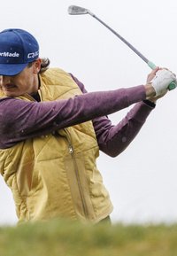 Žltá prešívaná vesta prehodená cez fialovú košeľu s dlhým rukávom, drží golfovú hrou, na hlave má modrú čiapku a na pravej ruke gripovú rukavicu.