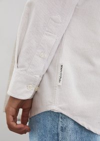 Camicia color crema a righe sottili con polsini abbottonati, con un'etichetta di stoffa sull'orlo che riporta "Marc O'Polo". Jeans blu denim in vista.