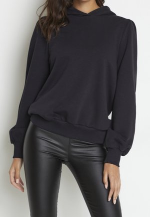 Femme portant un sweat à capuche noir avec des manches bouffantes et un pantalon noir brillant en simili cuir, debout devant un fond clair uni.