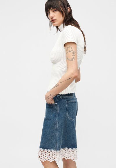 Γυναίκα με μακριά σκούρα μαλλιά φοράει ένα λευκό ριμπ κοντομάνικο τοπ και μια τζιν φούστα μέχρι το γόνατο με άσπρη δαντελένια διακόσμηση, στέκεται πλάγια.