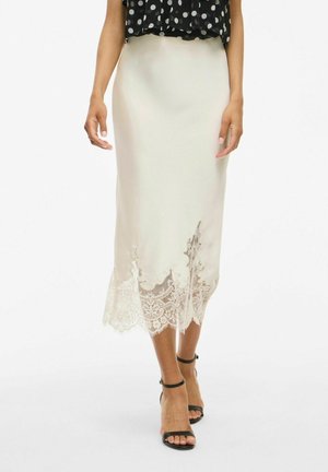 Jupe midi en soie crème avec un ourlet en dentelle à volants. Présente une texture lisse et une silhouette ajustée, associée à des escarpins noirs à lanières.