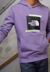 Sudadera con capucha morada y bolsillo frontal. Presenta un logo rectangular negro con texto blanco y acentos verdes. Textura de tela suave.