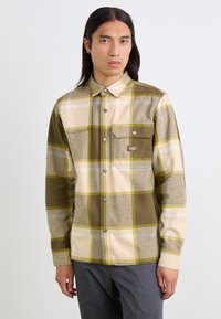 Chemise en flanelle à carreaux pour homme en vert olive, crème et gris. Détails en boutons argentés, poche poitrine et col classique. Texture douce.