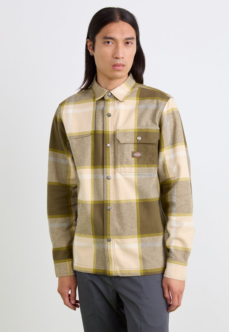 Chemise en flanelle à carreaux pour homme en vert olive, crème et gris. Détails en boutons argentés, poche poitrine et col classique. Texture douce.