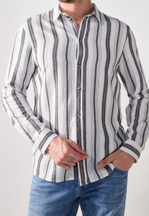 Mand iført afslappet langærmet skjorte med lodrette sorte, hvide og grå striber, kombineret med blå jeans, hånd justerer skjortens forside.