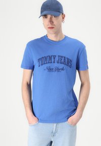 Mėlyna medvilninė marškinėliai trumpomis rankovėmis, su tamsiai mėlynu užrašu "TOMMY JEANS" ir "Niujorkas" spausdintu po juo. Šviesiai mėlyni džinsai ir suderinama kepurė.
