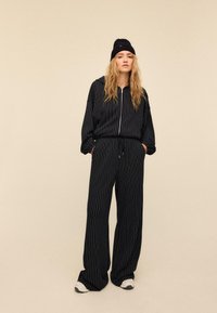 Černobílá proužkovaná bunda na zip a široké kalhoty, obojí z měkké, texturované látky. Kombinováno s černou čepicí a bílými teniskami.