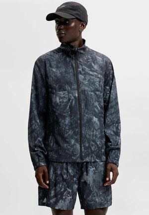 Personne noire portant une veste zippée noire et grise marbrée, un short assorti et une casquette noire, regardant vers la gauche sur un fond uni.