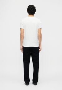 Άνδρας με σγουρά μαύρα μαλλιά στέκεται με την πλάτη στραμμένη φορώντας απλό λευκό κοντομάνικο μπλουζάκι και μαύρο παντελόνι μπροστά σε λευκό φόντο.