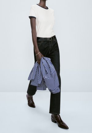Personne portant un t-shirt blanc avec des bordures noires, un jean noir et des bottes marron, tenant dans une main une chemise rayée bleue et blanche.