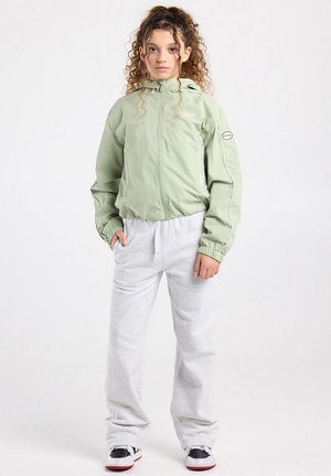 Jongere met krullend haar draagt een lichtgroene jas met capuchon, lichtgrijze joggingbroek en zwart-witte sneakers, staand tegen een witte achtergrond.