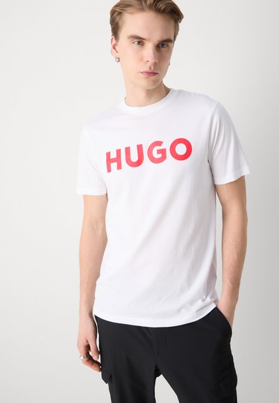 Бяла памучна тениска с ярко червено лого "HUGO" отпред, с кръгло деколте и къси ръкави, в комбинация с черни панталони.